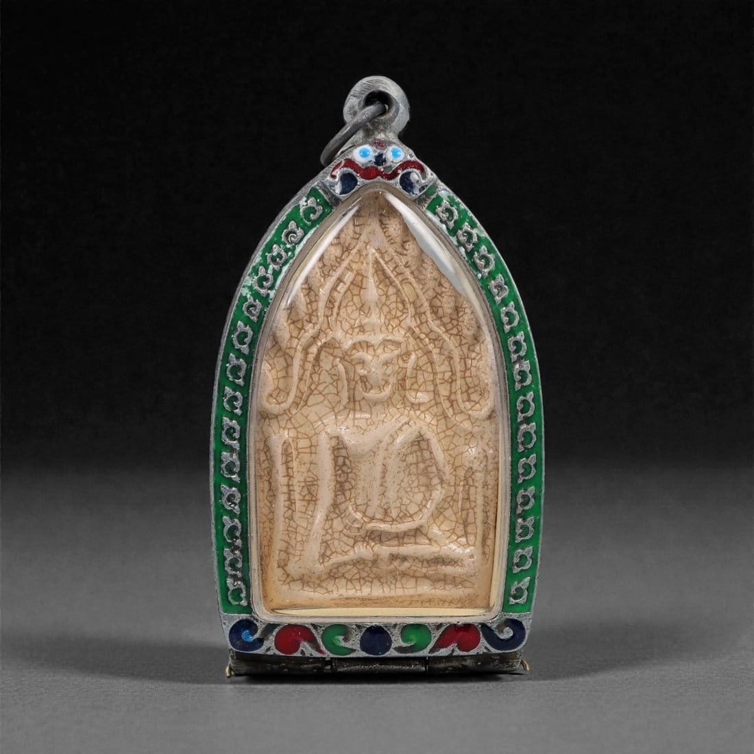 A Thai Buddhist Composite Amulet in Enameled Silvered Metal Frame, 20th Century, 52FN: The amulet is housed in an arched, tapering silvered metal frame with a suspension loop at the apex. The obverse frame is embellished with applied green, red, and blue enamel inlay within chased borde