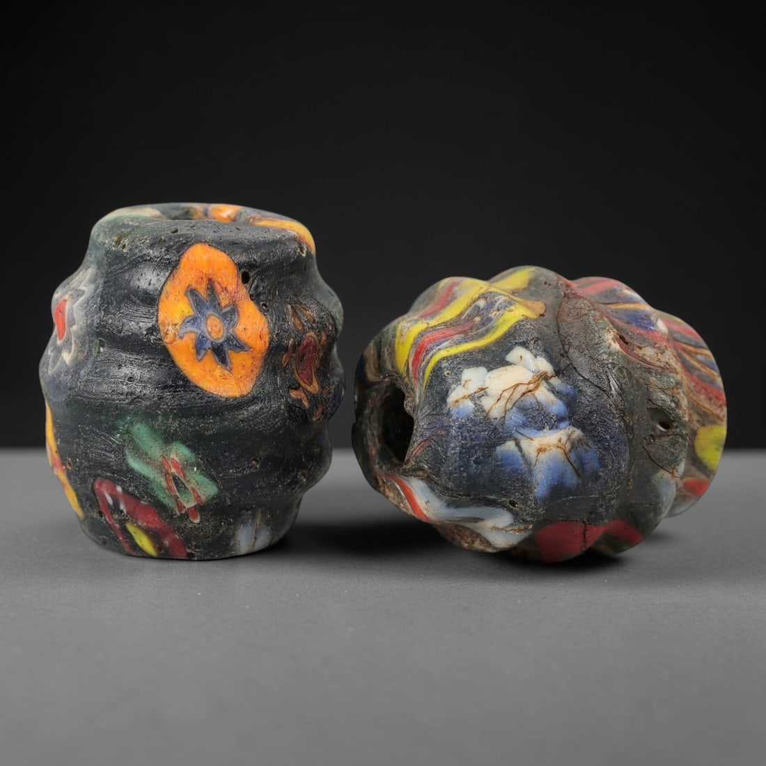 A Pair of Venetian Glass Trade Beads, 19th-20th Century, 77ZM: Formed as lobed, barrel-shaped beads, each is crafted from dark translucent glass with a central perforation for suspension. The surface is decorated with multiple polychrome floral and abstract motif