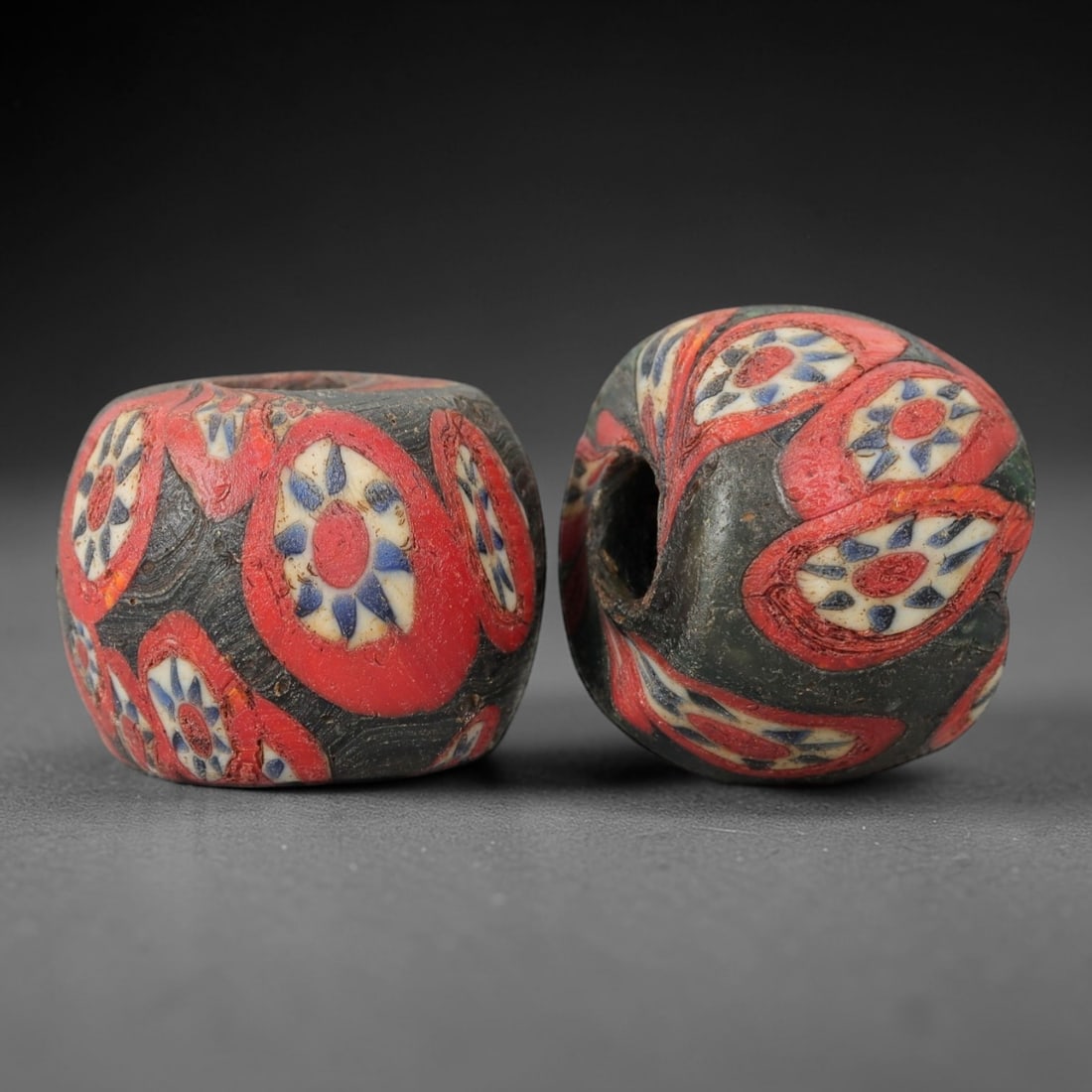 A Pair of Venetian Glass Trade Beads, 19th-20th Century, 64ZM: Formed as barrel-shaped beads, each is crafted from dark translucent glass with a central perforation for suspension. The surface is decorated with multiple polychrome floral motifs, each featuring a
