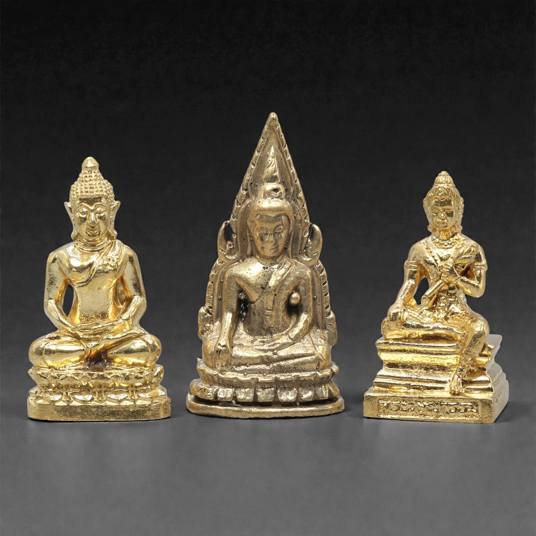 A Set of Three Thai Brass Buddha and Deity Figures, 20th Century, 73ZG: Cast from brass with a bright, gilded finish, the set comprises three distinct seated figures. The left figure is a Buddha seated in dhyana mudra, mounted on a detailed lotus base. The central figure