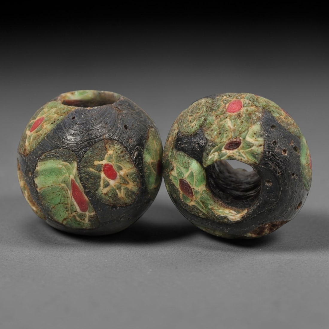 A Pair of Venetian Millefiori Glass Trade Beads, 19th-20th Century, 97ZM: Formed as spherical beads, each is crafted from translucent dark glass with a central perforation for suspension, showcasing the intricate millefiori technique. The surface is adorned with repeating p