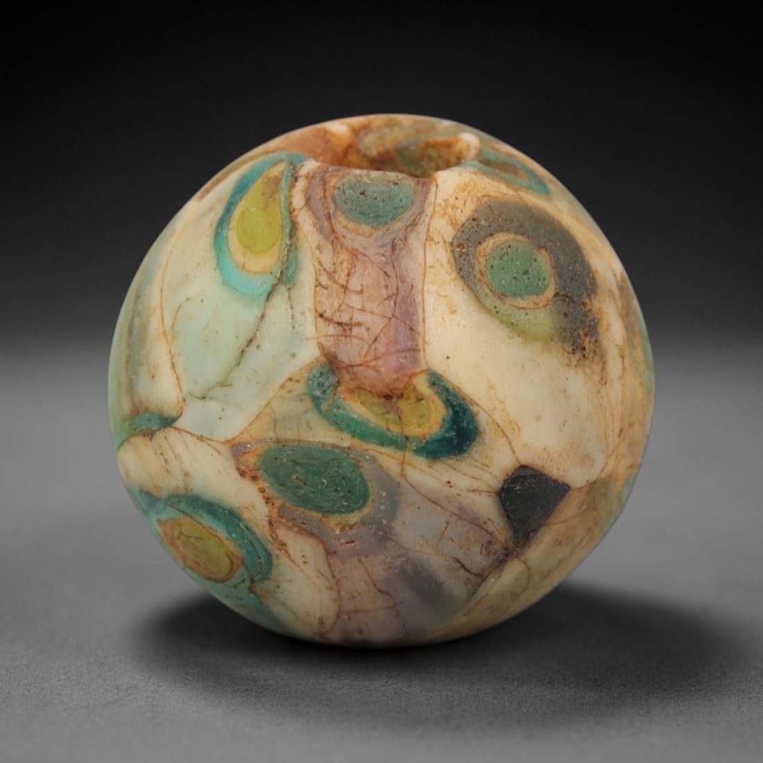 A Venetian Millefiori Glass Bead, 19th-20th Century, 26ZM: Fabricated from translucent and opaque glass, the bead is spherical with a central perforation for suspension. The surface displays a millefiori pattern of circular inlays in green, yellow, and teal,