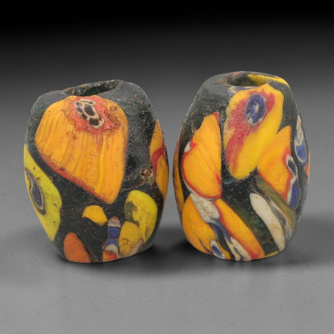 A Pair of Venetian Millefiori Glass Beads, 19th-20th Century, 71AD: Crafted from opaque dark green glass, each bead is formed as an elongated bicone with a central drilled perforation. The surface is decorated with characteristic millefiori canes, featuring large abst