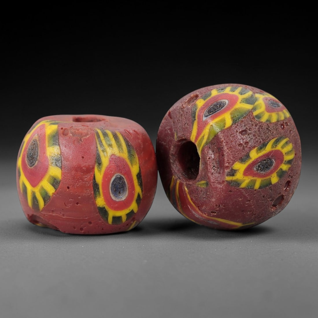 A Pair of Venetian Glass Trade Beads, 19th-20th Century, 81ZM: Formed as barrel-shaped beads, each is crafted from opaque red glass with a central perforation for suspension. The surface is decorated with multiple polychrome eye motifs, each featuring a dark core
