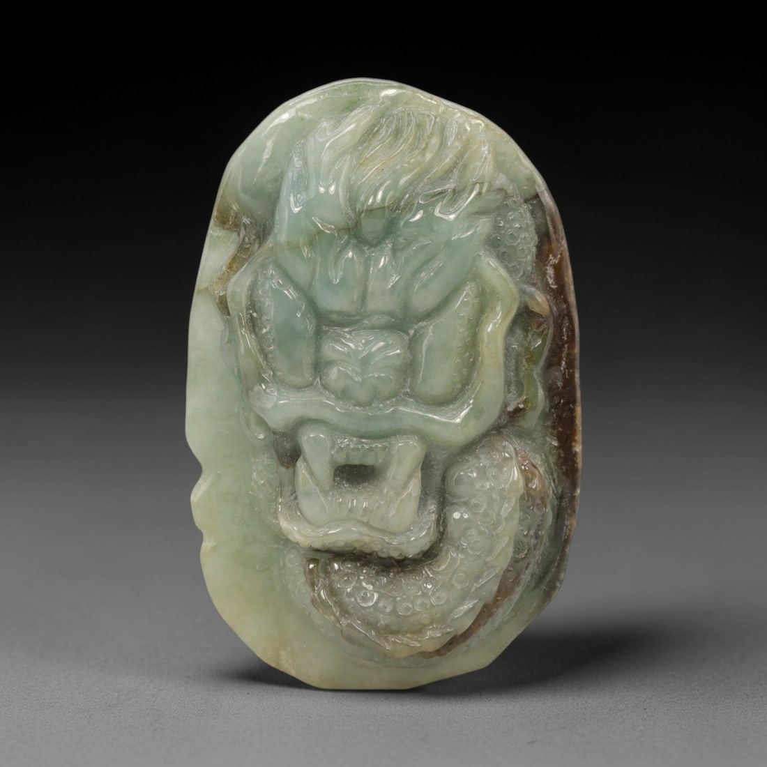 A Chinese Jadeite Jade Dragon Pendant, 20th Century, 8ZL: Carved from pale green jadeite with natural brownish inclusions along the edges, the object is formed as an irregularly oval pendant. The front is decorated in high relief with a dragon head, featurin