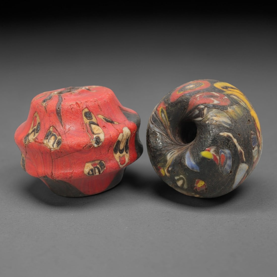 A Pair of Venetian Glass Trade Beads, 19th-20th Century, 87ZM: Formed as lobed, barrel-shaped and spherical beads, each is crafted from opaque red and dark translucent glass respectively, with a central perforation for suspension. The surface is decorated with mu