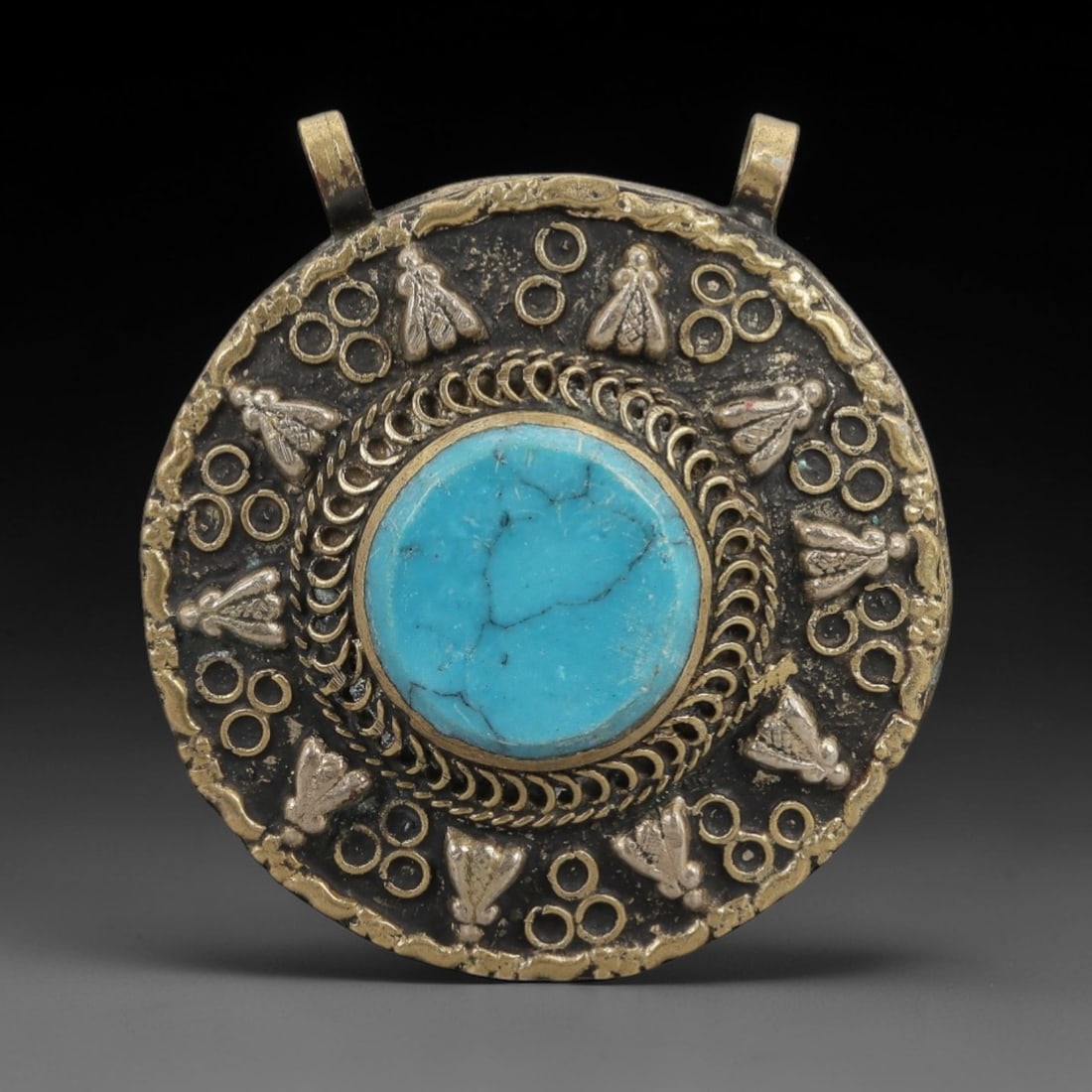 A Tibetan Silver and Turquoise Amulet Pendant, 20th Century, 88ZL: Crafted from a silver and brass alloy with a dark, aged patina, the pendant is of circular form, centered by a large, cabochon-cut turquoise stone with prominent natural veining. The stone is set with