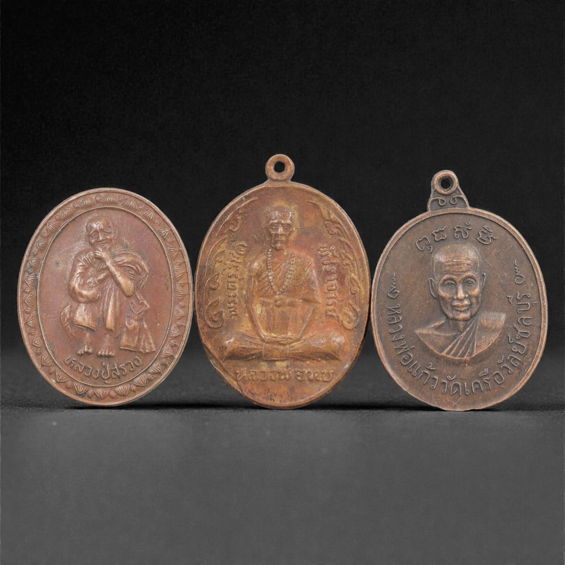 Three Thai Copper Alloy Devotional Amulets, 20th Century, 69FN: Cast from copper alloy with a warm brown patina, the group comprises three oval-shaped devotional amulets, each with a suspension loop at the apex for wear. The first amulet depicts a seated figure wi