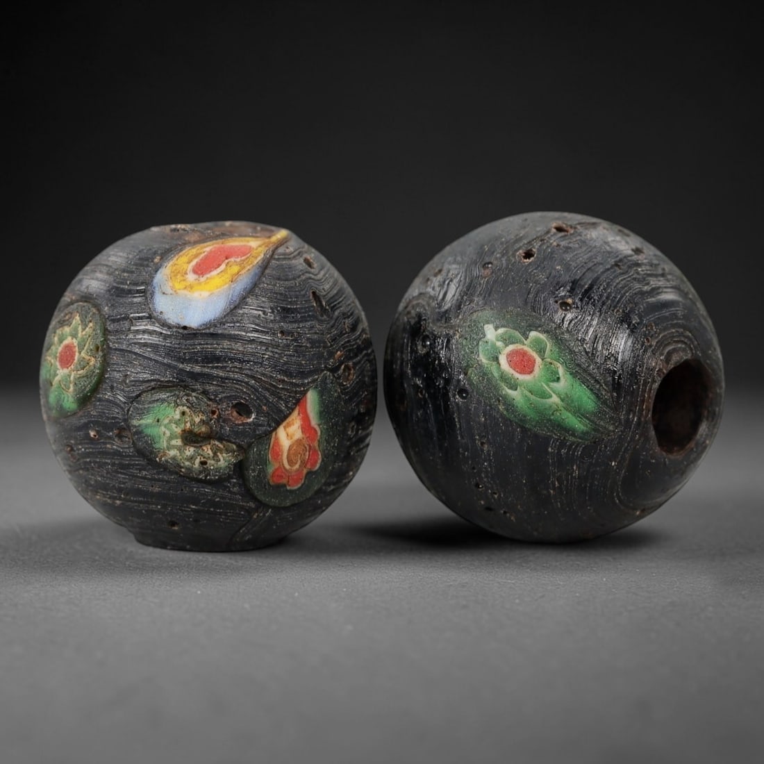 A Pair of Venetian Glass Trade Beads, 19th-20th Century, 82ZM: Formed as spherical beads, each is crafted from dark translucent glass with a central perforation for suspension. The surface is decorated with multiple polychrome floral and eye motifs, featuring red