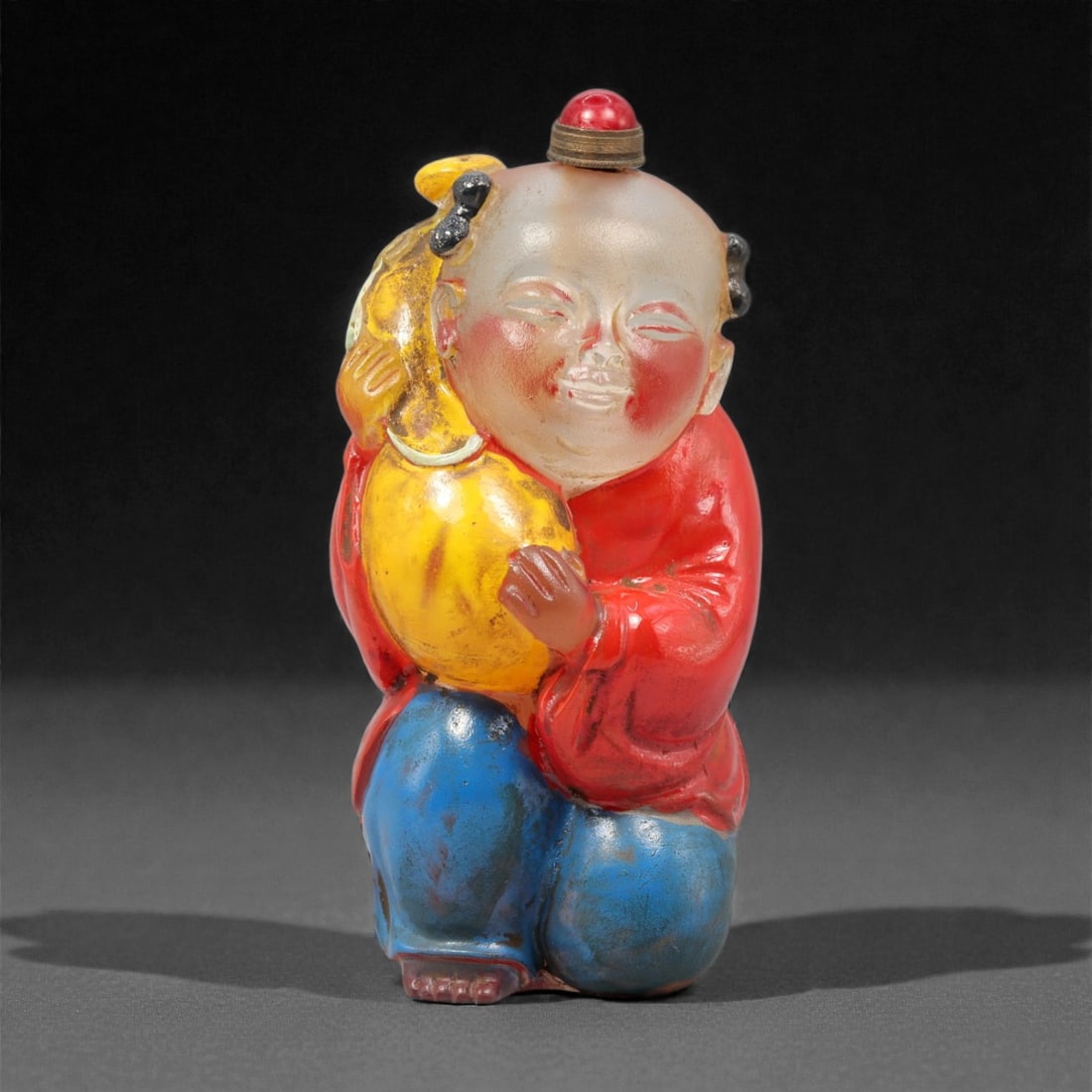 A Chinese Figural Glass Snuff Bottle, 20th Century, 64YC: Molded in the form of a smiling child kneeling on one knee, this figural snuff bottle is crafted from translucent and opaque glass and finished in bright polychrome enamels. The child is shown wearing