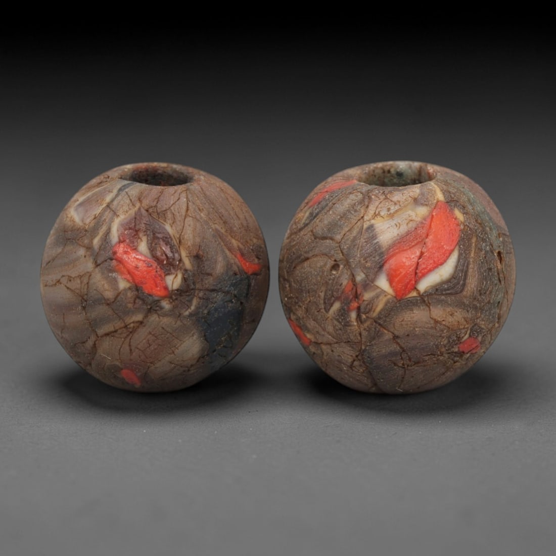 A Pair of Venetian Millefiori Glass Beads, 19th-20th Century, 59AD: Crafted from translucent brownish glass with dark inclusions, each bead is formed as a sphere with a central drilled perforation. The surface displays marbled swirled patterns in red, white, and dark