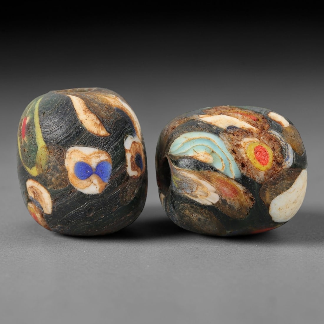 A Pair of Venetian Glass Trade Beads, 19th-20th Century, 84ZM: Formed as barrel-shaped beads, each is crafted from dark translucent glass with a central perforation for suspension. The surface displays a marvered, swirled pattern of opaque colored inclusions, inc