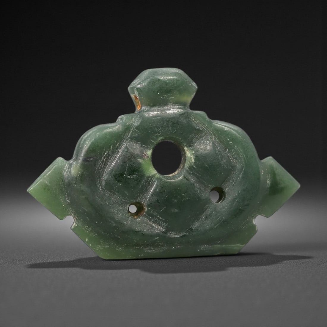An Antique Islamic Jade Amulet, 10th-14th Century CE, 76FL: Carved from translucent green jade with natural tonal variation and minor surface scratches, the amulet is formed as a symmetrical, shield-shaped plaque. A large central circular perforation is flanke