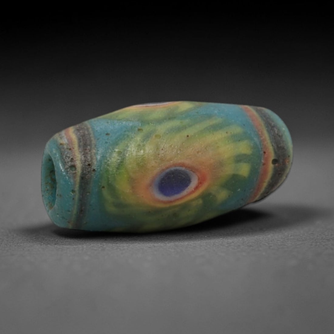 A Sino-Himalayan Glass Eye Bead, 18th-19th Century, 28ZN: Formed from translucent blue-green glass with swirling yellow and pale green filamentary inclusions characteristic of mosaic glass working, the bead is barrel-shaped with a central perforation for sus