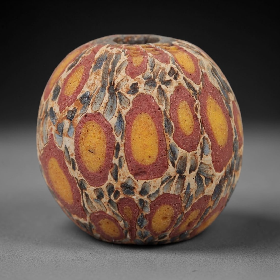 Ancient Islamic Glass Bead - Eye Mosaic Amulet Bead, 14FW: This spherical bead is crafted from layered mosaic glass, featuring repeating eye-shaped motifs with yellow centers, red outer rings, and intricate blue-and-cream matrix patterns, characteristic of me