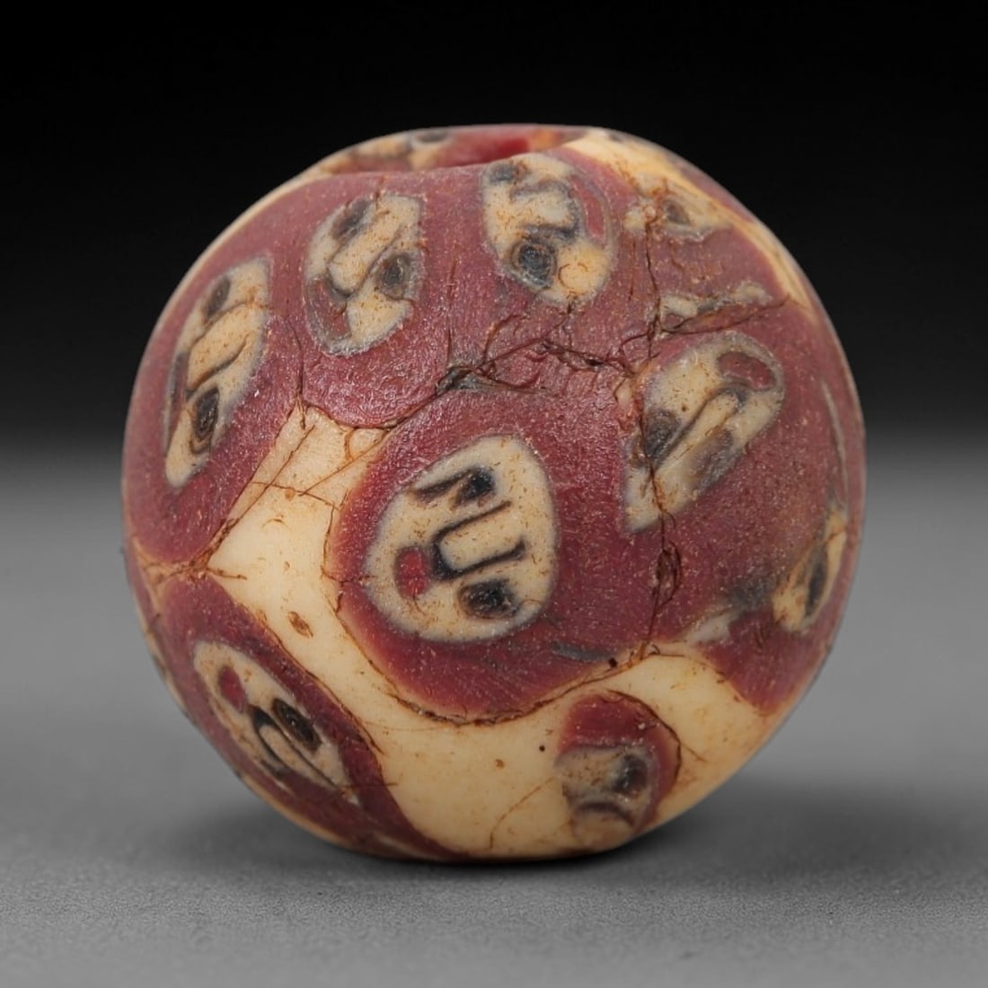 Ancient Glass Face Bead - Roman/Egyptian Amulet Bead, 50FW: This spherical bead is crafted from layered glass, featuring a creamy white base overlaid with deep red sections, each punctuated by stylized face motifs in black and red. The faces are rendered with