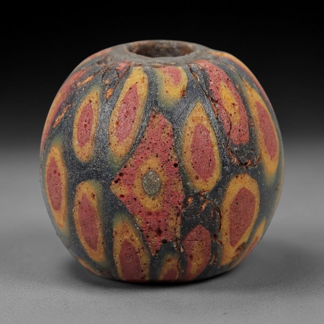 Ancient Glass Eye Bead - Roman/Egyptian Mosaic Amulet Bead, 33FW: This spherical bead is crafted from layered mosaic glass, featuring a dark blue base overlaid with repeating eye-shaped motifs in red, yellow, and tan, each centered with a cool-toned circular core, e