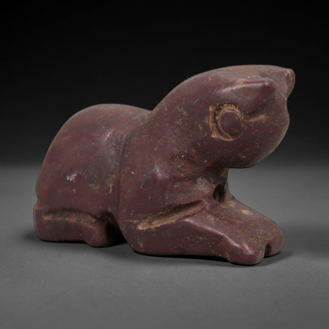 Ancient Dark Red Stone Recumbent Feline - Near Eastern Amulet, 13AE: Carved from deep reddish-purple stone with natural granular texture, this amulet depicts a recumbent feline in a compact, stylized form. The creature is rendered in a prone resting posture, with simpl
