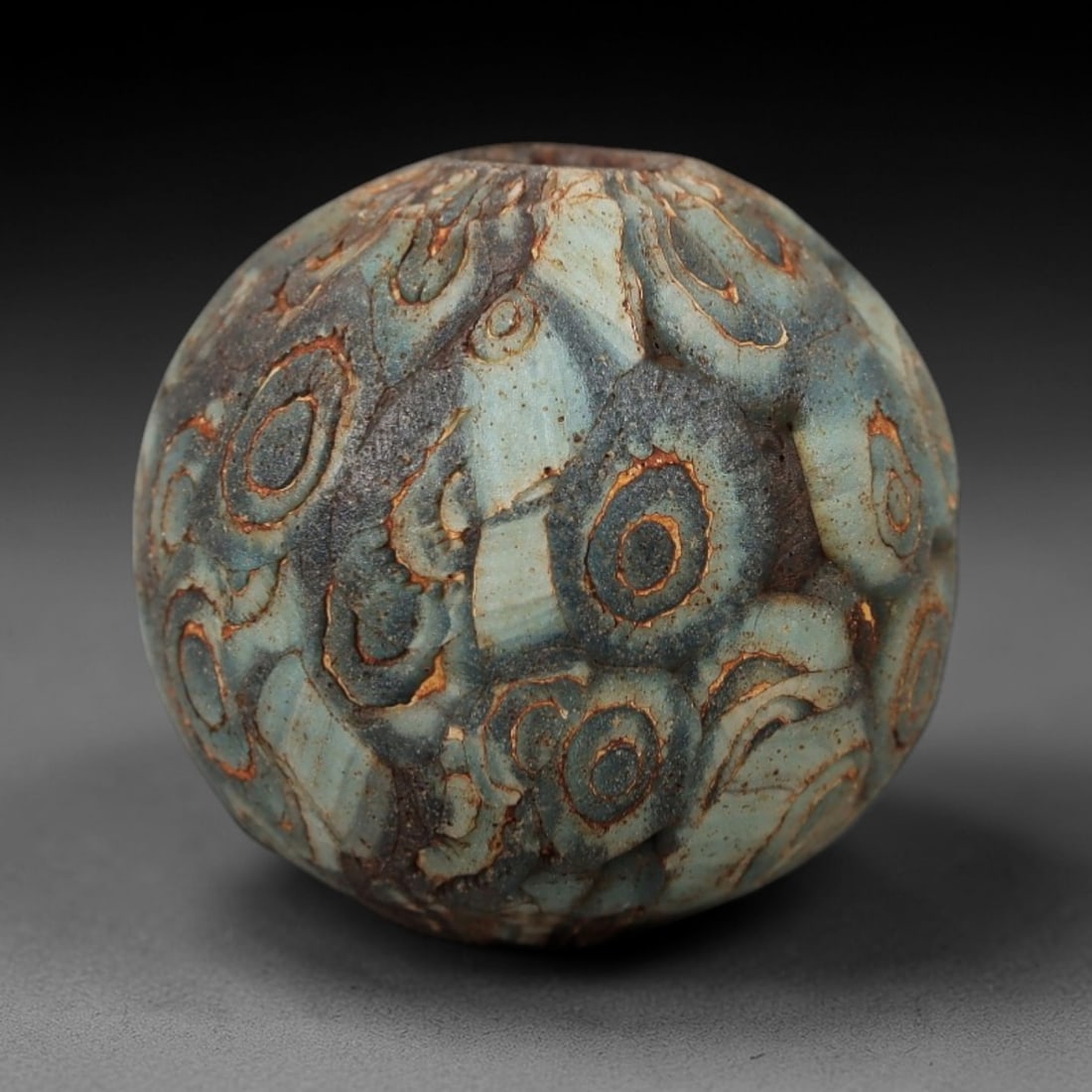 Ancient Roman/Egyptian Glass Bead - Eye Mosaic Amulet Bead, 23FW: This spherical bead is crafted from layered pale turquoise glass, adorned with dense repeating eye-shaped motifs-each with a dark center, pale blue ring, and rust-colored outline-evoking the protectiv