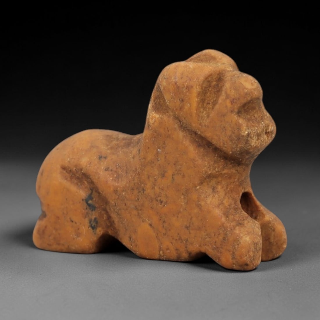 Ancient Tan Stone Recumbent Feline - Near Eastern Amulet, 87AD: Carved from warm tan stone with natural granular texture and subtle dark mineral inclusions, this amulet depicts a recumbent feline in a compact, stylized form. The creature is rendered in a prone res