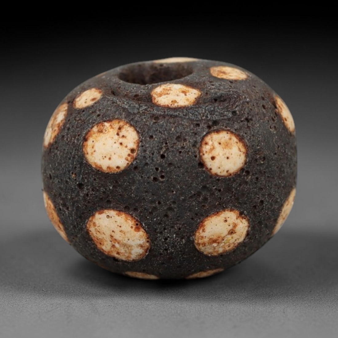 Ancient Glass Spotted Bead - Roman/Egyptian Amulet Bead, 24FW: This spherical bead is crafted from porous dark glass, adorned with evenly spaced circular white inlays edged with warm orange-brown patina, characteristic of Roman and Egyptian glassworking tradition