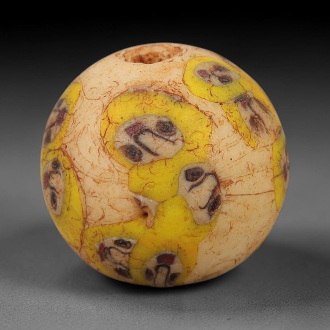Ancient Roman/Egyptian Glass Bead - Face Mosaic Amulet Bead, 53FW: This spherical bead is crafted from creamy ivory glass, adorned with repeating stylized face motifs set within yellow medallions, each featuring simplified eyes, a curved nose, and a red mouth-evoking