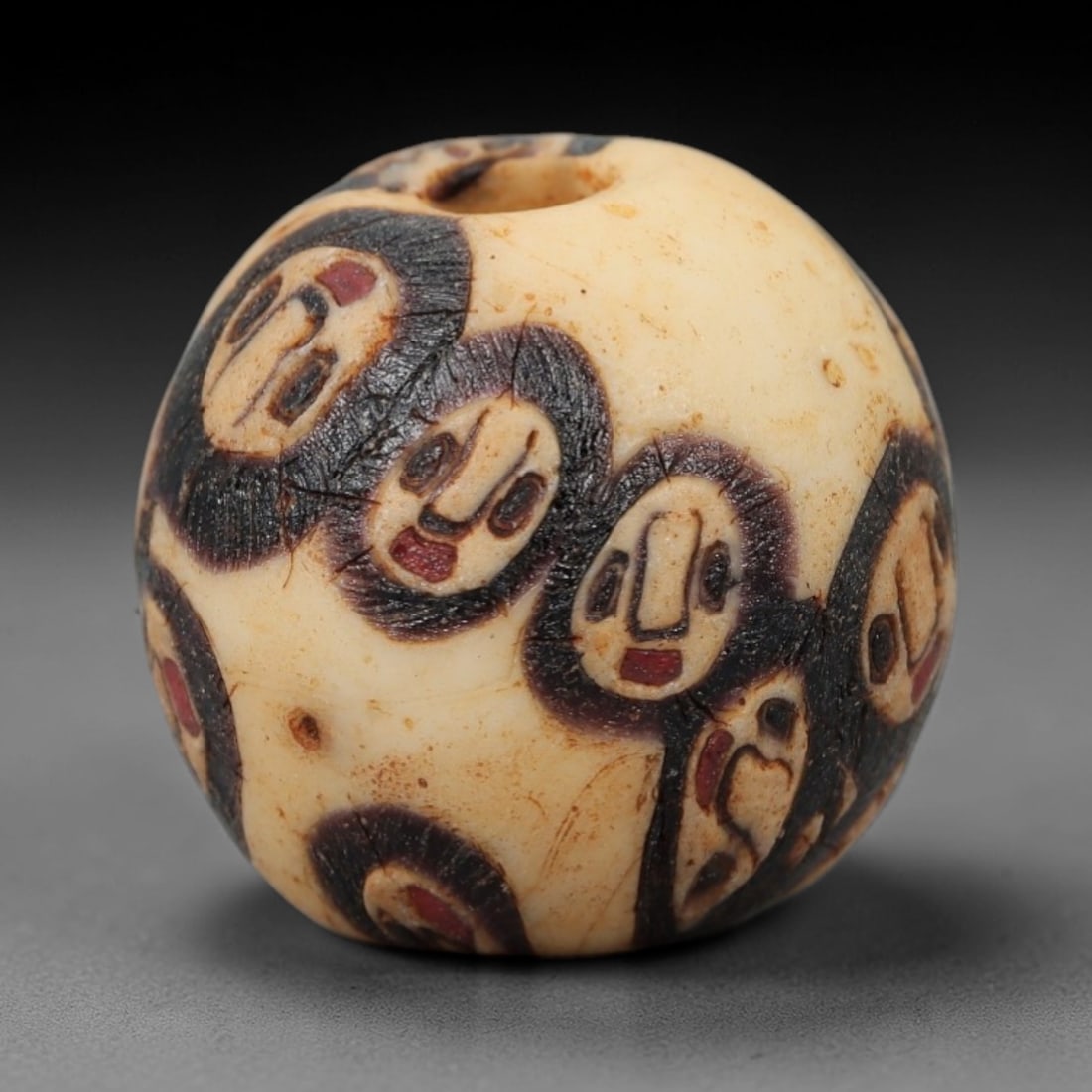 Ancient Glass Face Bead - Roman/Egyptian Mosaic Amulet Bead, 34FW: This spherical bead is crafted from creamy ivory glass, adorned with repeating stylized face motifs in black and red, each featuring simplified eyes, a defined nose, and a red mouth, evoking the prote