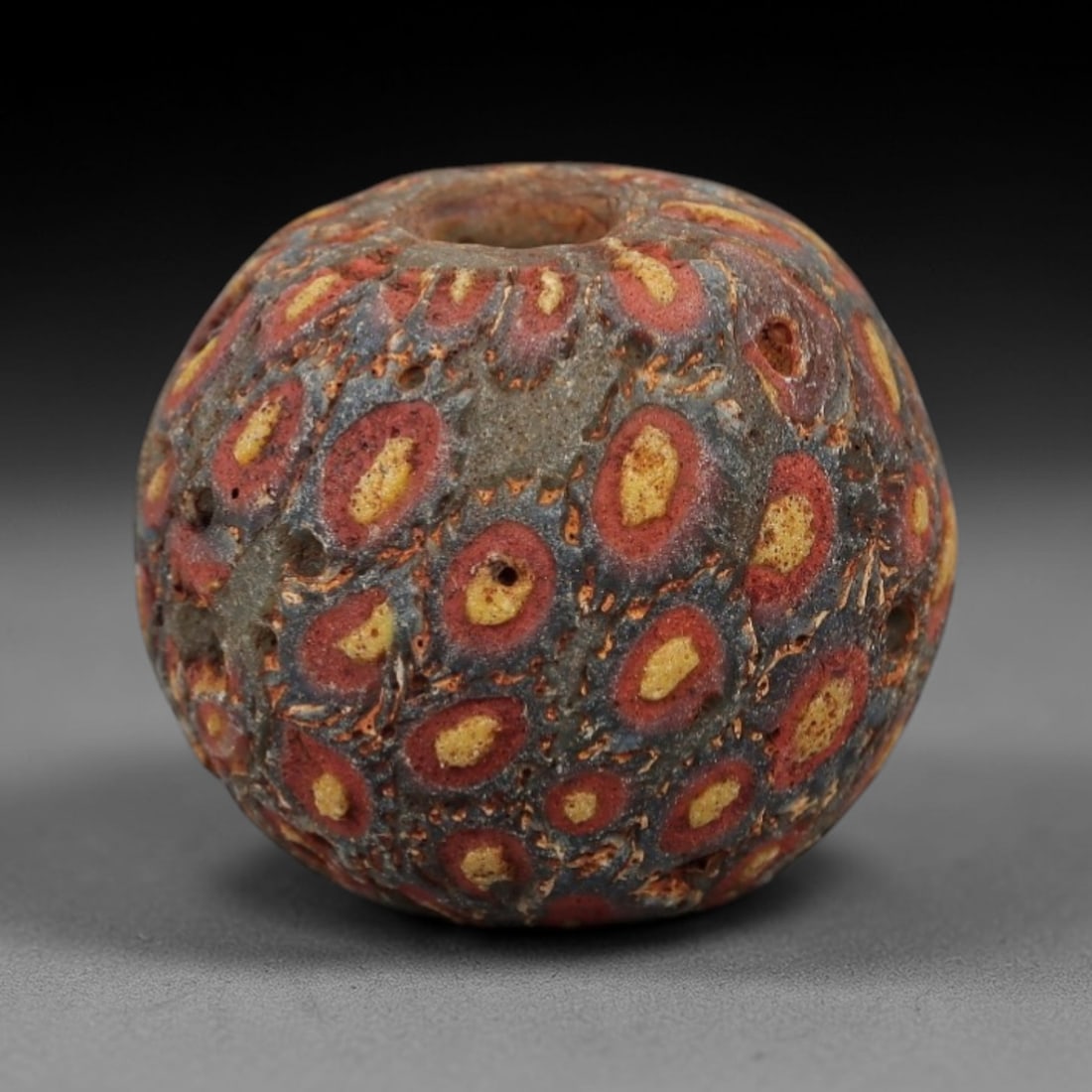 Ancient Glass Eye Bead - Roman/Egyptian Mosaic Amulet Bead, 21FW: This spherical bead is crafted from layered mosaic glass, featuring a muted grey-blue base overlaid with repeating eye-shaped motifs in red and yellow, each centered with a warm yellow core, evoking t