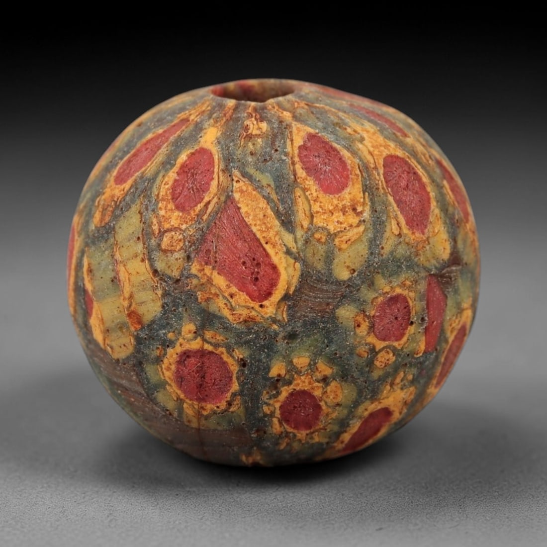 Ancient Glass Eye Bead - Roman/Egyptian Mosaic Amulet Bead, 43FW: This spherical bead is crafted from layered mosaic glass, featuring a muted green-grey base overlaid with repeating eye-shaped motifs in red and yellow, each centered with a vivid red core, evoking th