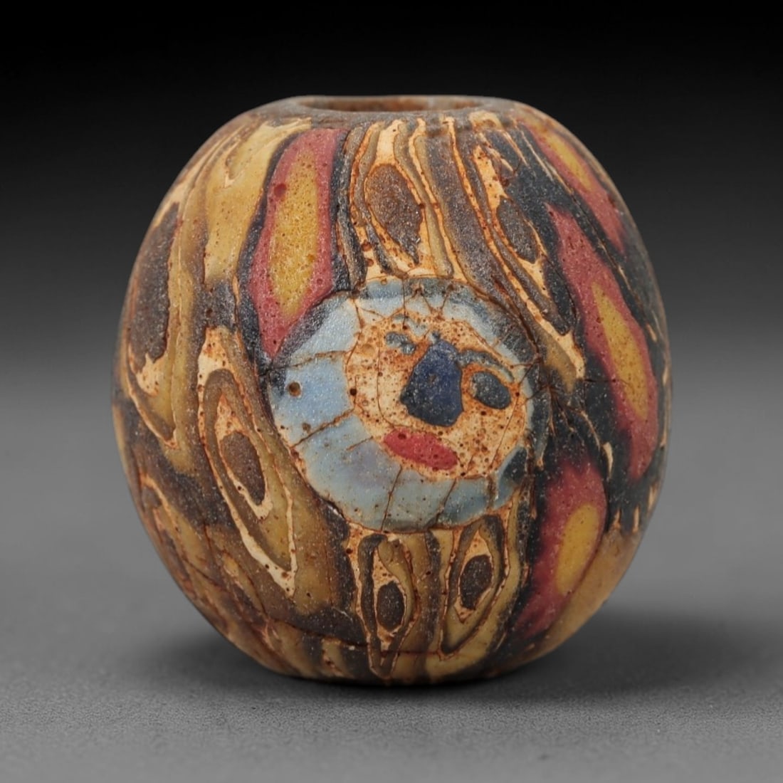 Ancient Glass Face Bead - Roman/Egyptian Mosaic Amulet, 10FW: This ovoid bead is crafted from layered mosaic glass, featuring a warm yellow-brown base overlaid with swirling red, black, and white linear patterns, centered on a prominent stylized face motif. The