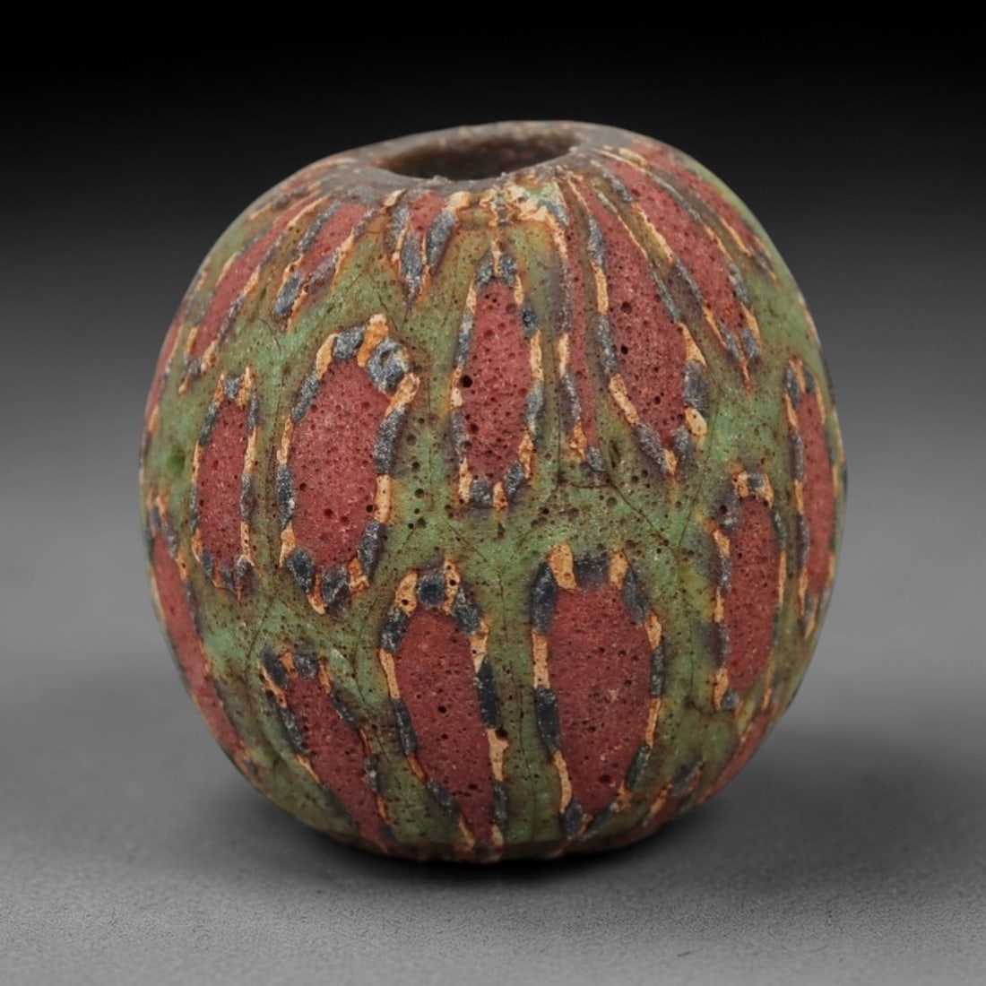Ancient Islamic Glass Bead - Geometric Mosaic Amulet Bead, 30FW: This spherical bead is crafted from porous red and green layered glass, featuring repeating geometric oval motifs outlined in blue and cream, characteristic of medieval Islamic glassworking traditions