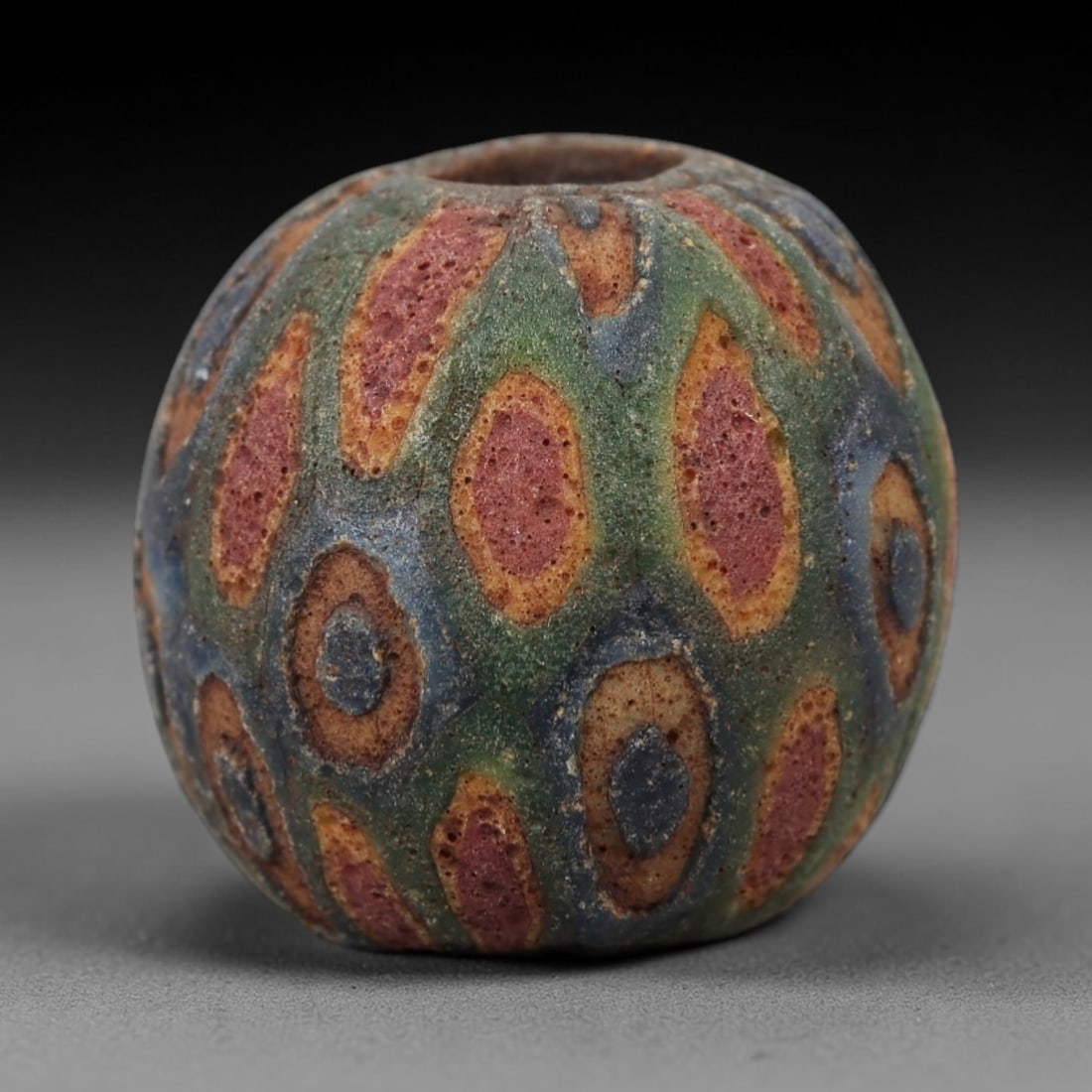 Ancient Islamic Glass Bead - Eye Mosaic Amulet Bead, 12FW: This spherical bead is crafted from layered porous glass, featuring repeating eye-shaped motifs in red, yellow, and deep blue against a muted green base-characteristic of medieval Islamic glassworking