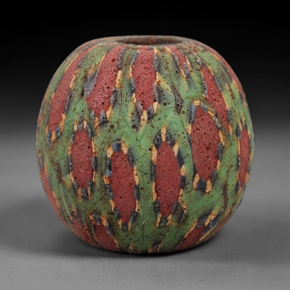 Ancient Islamic Glass Bead - Geometric Mosaic Amulet Bead, 32FW: This spherical bead is crafted from porous red and green layered glass, featuring repeating geometric oval motifs outlined in blue and cream, characteristic of medieval Islamic glassworking traditions