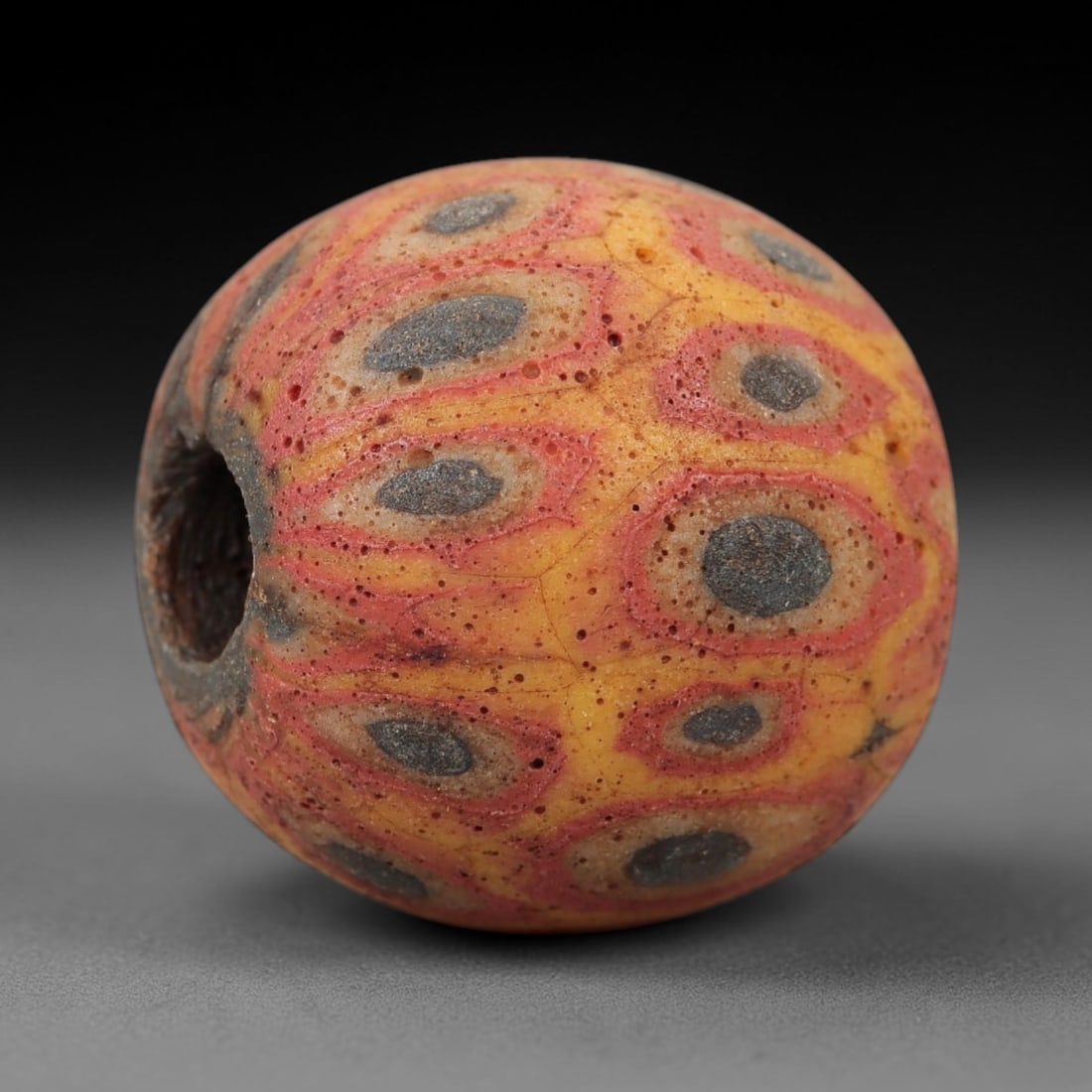 Ancient Glass Eye Bead - Roman Mosaic Amulet, 9FW: Crafted from opaque yellow and red glass with dark blue inclusions, this spherical bead employs the mosaic glass technique, where colored glass canes are fused and sliced to form repeating eye-like mo