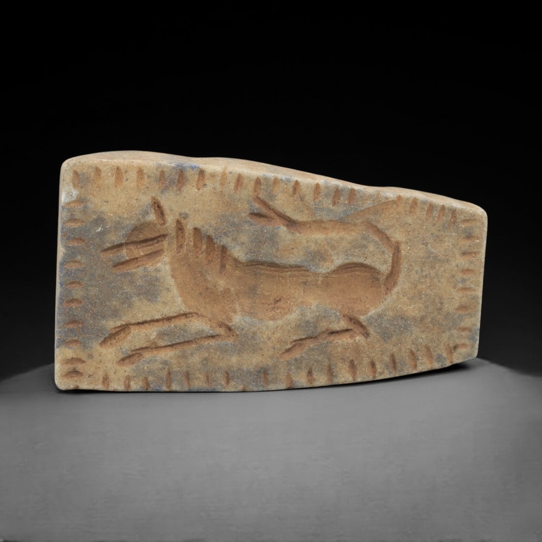 Ancient Lapis Plaque - Near Eastern Animal Figurine, 90FW: Carved from a speckled, earth-toned lapis lazuli plaque, this artifact depicts a leaping quadrupedal animal with incised lines defining its limbs, mane, and tail, framed by a border of punctuated dot