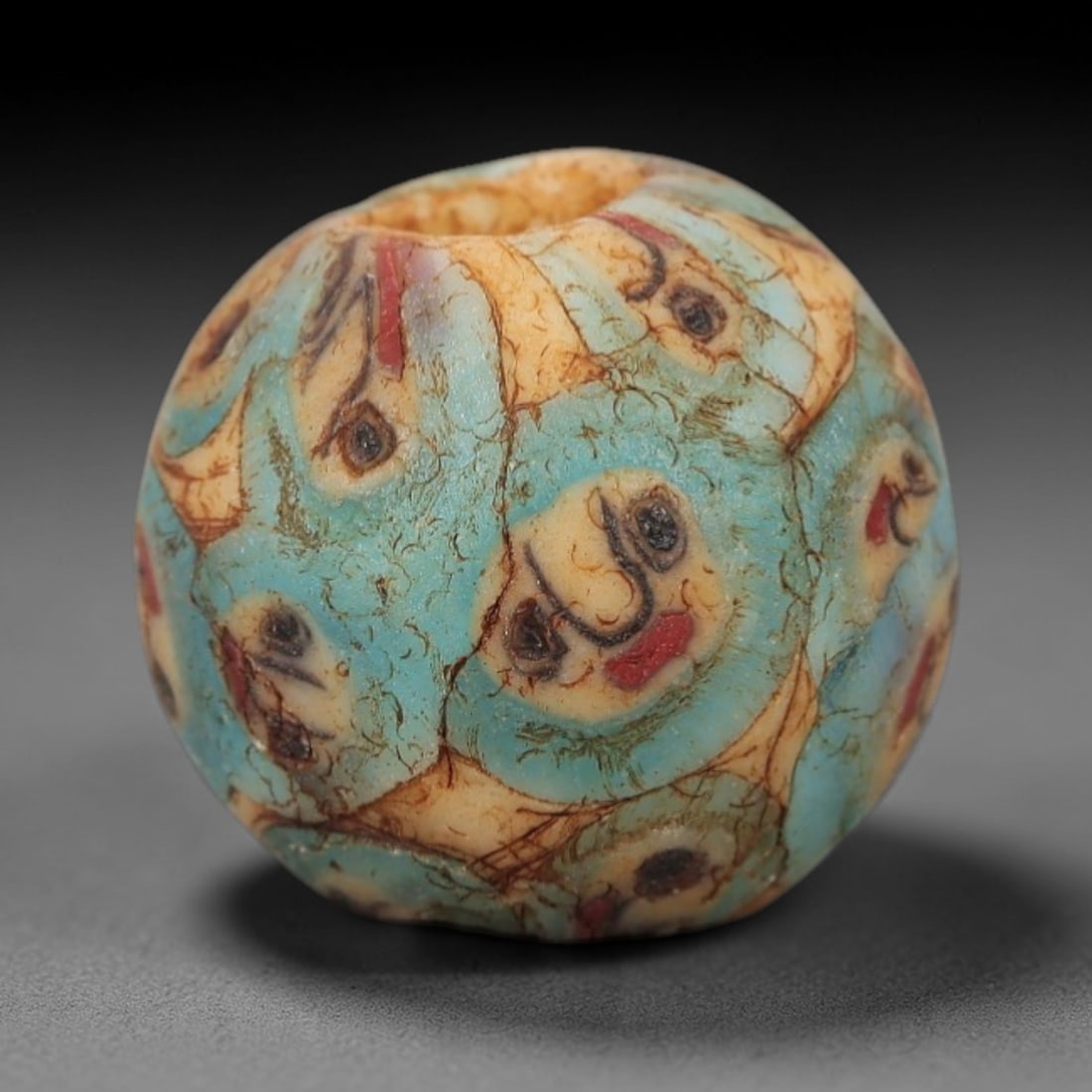 Ancient Glass Face Bead - Roman/Egyptian Mosaic Amulet Bead, 4FW: This spherical bead is crafted from translucent turquoise glass, adorned with repeating stylized face motifs in beige, black, and red, each featuring simplified eyes, a curved nose, and a red mouth, e