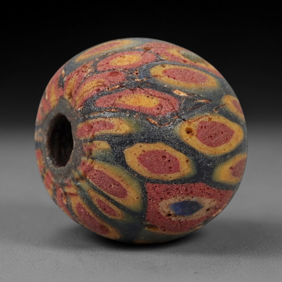 Ancient Glass Eye Bead - Roman/Egyptian Mosaic Bead, 37FW: This spherical bead is crafted from layered mosaic glass, featuring a dark blue base overlaid with repeating eye-shaped motifs in red, yellow, and a single blue central dot, evoking the protective "ey