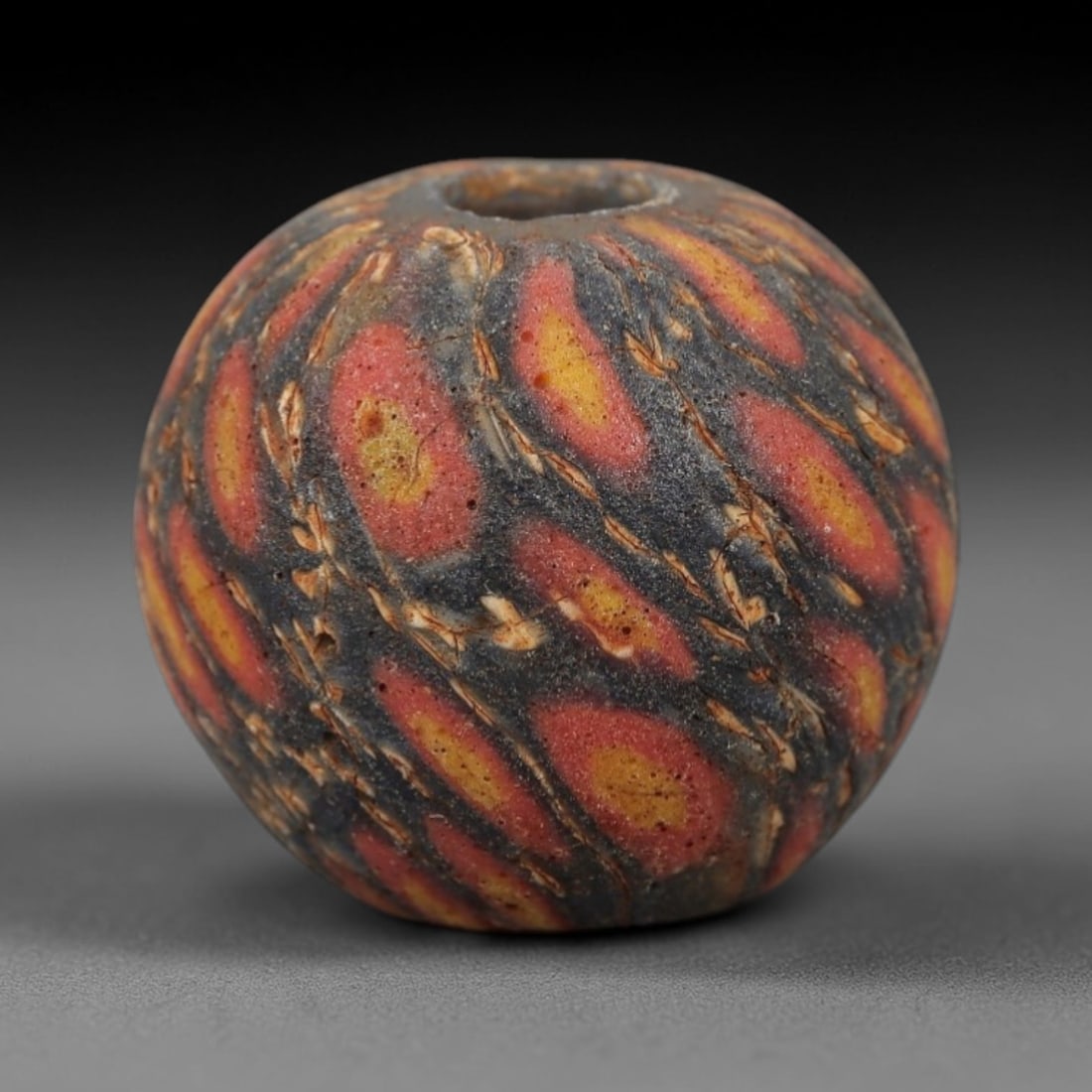 Ancient Islamic Glass Bead - Flame Mosaic Amulet Bead, 59FW: This spherical bead is crafted from layered porous glass, featuring repeating flame-shaped motifs in red and yellow against a deep indigo base-characteristic of medieval Islamic glassworking tradition