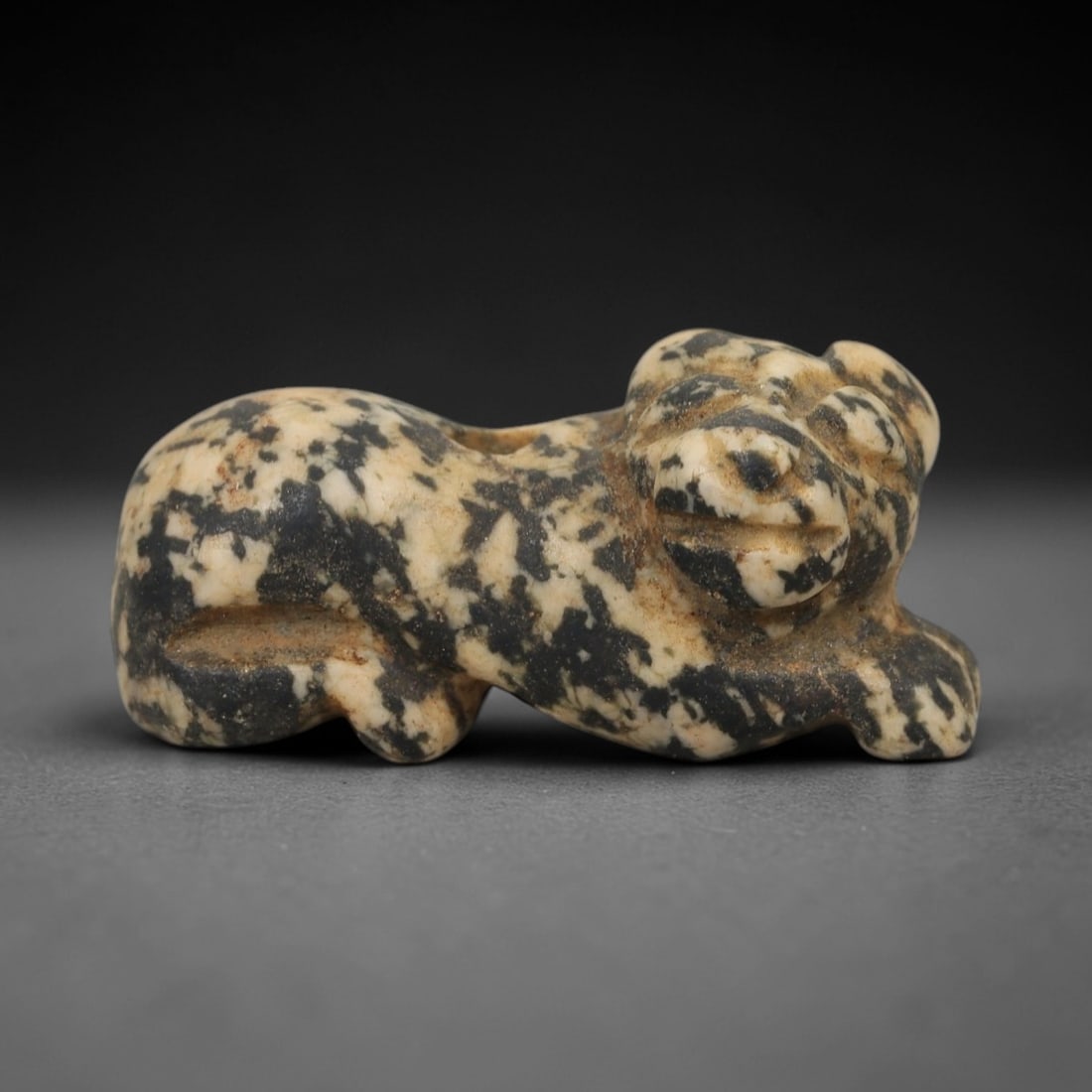 Ancient Dappled Stone Recumbent Bovid - Near Eastern Amulet, 11AE: Carved from dappled black-and-white stone, this amulet depicts a recumbent bovid in a compact, stylized form. The creature is rendered in a crouched, resting posture, with simplified facial features a