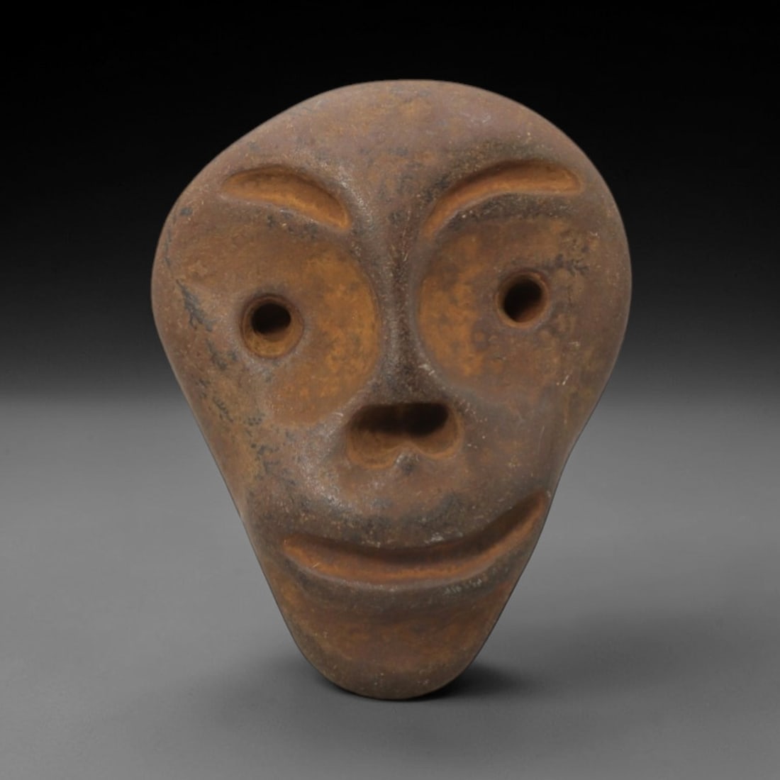 Ancient Stone Maskette - Medieval African Tribal Figurine, 76FW: Carved from a coarse, earthy stone, this medieval African tribal maskette presents a stylized anthropomorphic face with rounded, recessed eye sockets, a heart-shaped nasal aperture, arched brows, and
