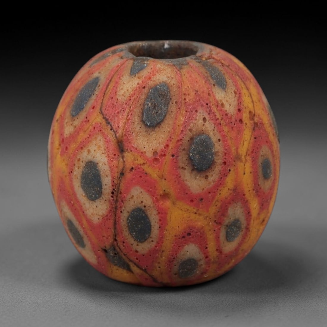 Ancient Glass Eye Bead - Roman Mosaic Amulet, 27FW: Crafted from opaque orange and red glass with dark blue inclusions, this spherical bead utilizes the mosaic glass technique, where colored glass canes are fused and sliced to form repeating eye-like m