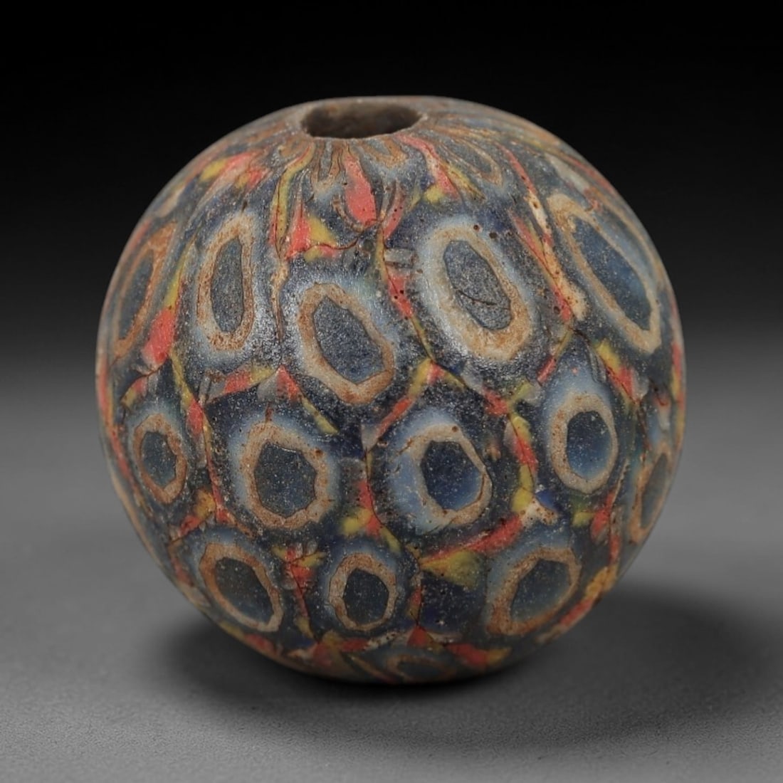 Ancient Roman/Egyptian Glass Bead - Eye Mosaic Amulet Bead, 26FW: This spherical bead is crafted from deep indigo glass, adorned with repeating eye-shaped motifs-each with a dark center, creamy white ring, and vibrant red-yellow accents-evoking the protective eye am
