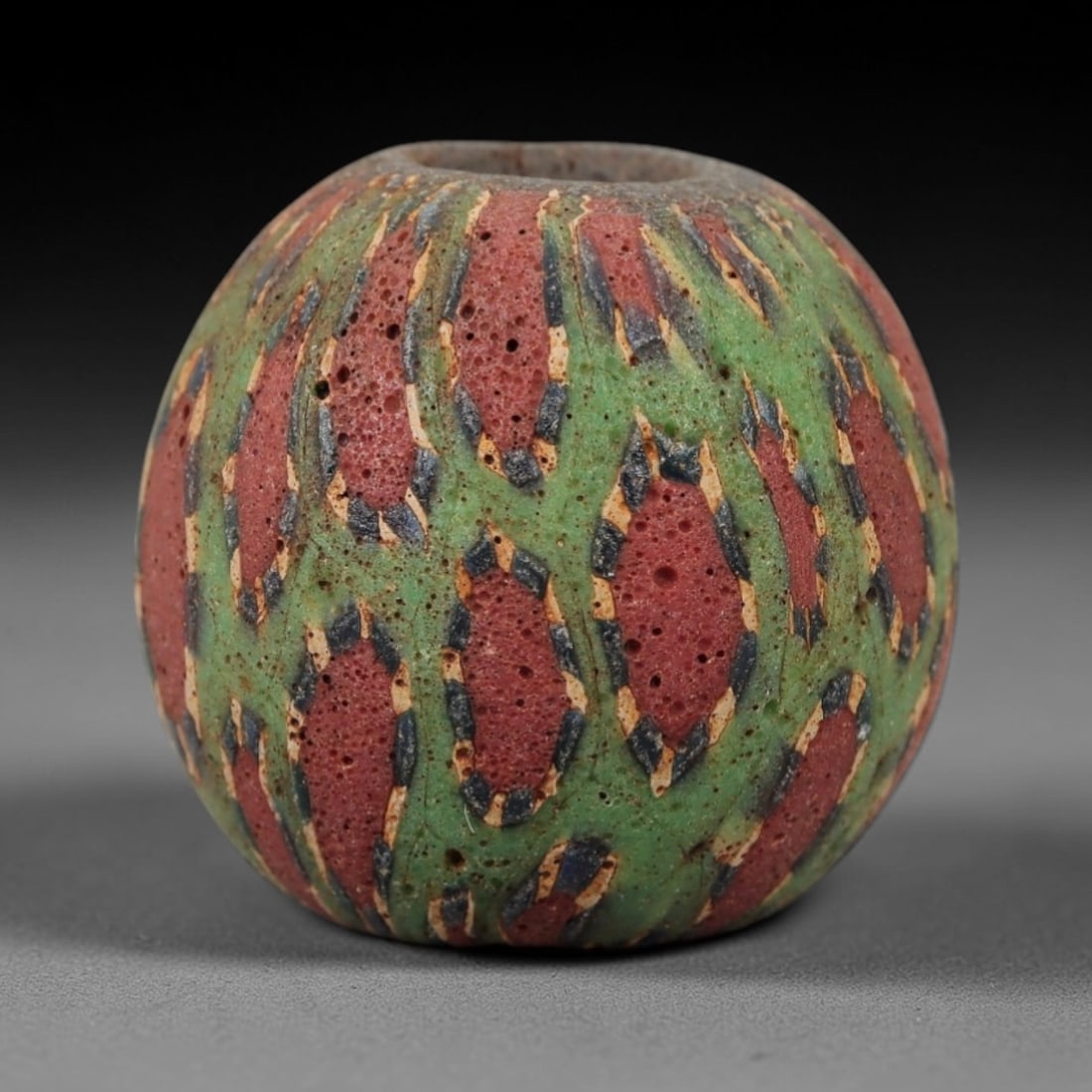 Ancient Islamic Glass Bead - Geometric Mosaic Amulet Bead, 40FW: This spherical bead is crafted from porous red and green layered glass, featuring repeating geometric oval motifs outlined in blue and cream, characteristic of medieval Islamic glassworking traditions