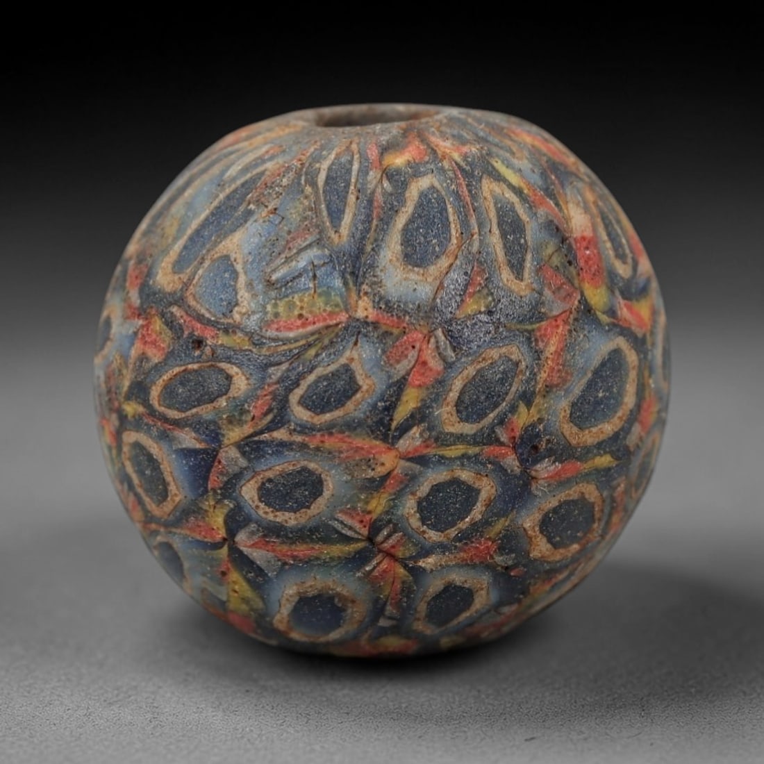 Ancient Roman/Egyptian Glass Bead - Eye Mosaic Amulet Bead, 15FW: This spherical bead is crafted from deep indigo glass, adorned with a dense array of eye-shaped motifs-each with a dark center, creamy white ring, and vibrant red-yellow accents-evoking the protective