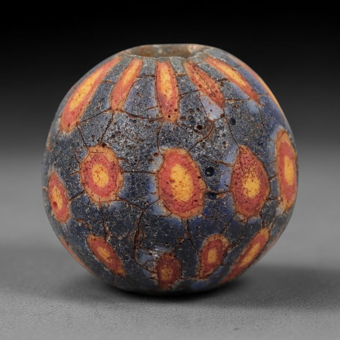 Ancient Glass Eye Bead - Roman Mosaic Amulet, 42FW: Crafted from translucent dark blue glass with opaque red and yellow inclusions, this spherical bead employs the mosaic glass technique, where colored glass canes are fused and sliced to form repeating