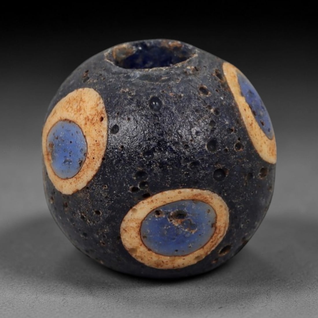 Ancient Roman/Egyptian Glass Bead - Eye Mosaic Amulet Bead, 49FW: This spherical bead is crafted from deep indigo glass, adorned with repeating eye-shaped motifs-each with a light blue center and creamy tan ring-evoking the protective eye amulet traditions of ancien