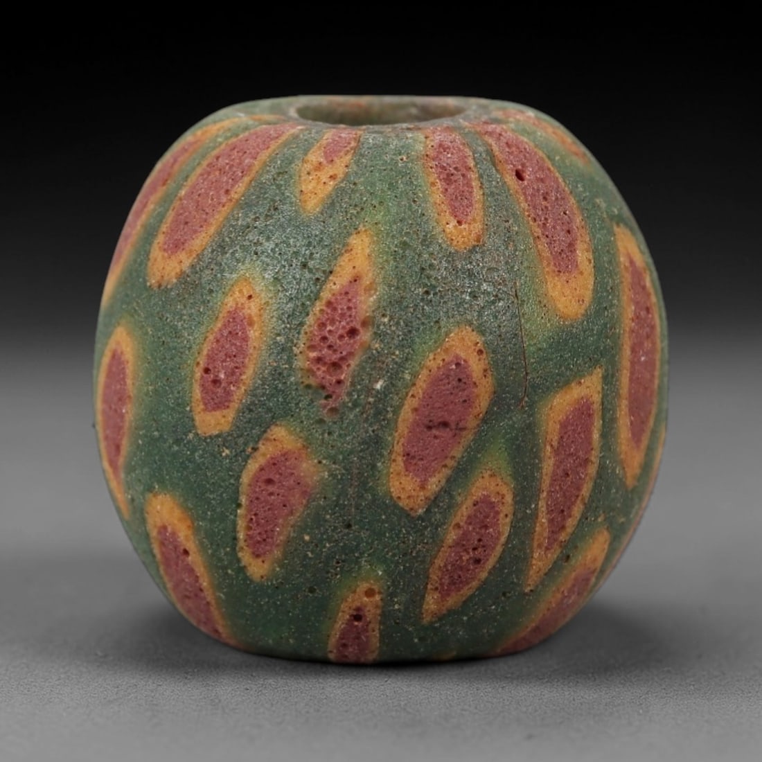 Ancient Islamic Glass Bead - Teardrop Pattern Amulet Bead, 8FW: This spherical bead is crafted from porous green glass, adorned with repeating teardrop-shaped motifs in red and yellow, characteristic of medieval Islamic glassworking traditions. A central axial hol