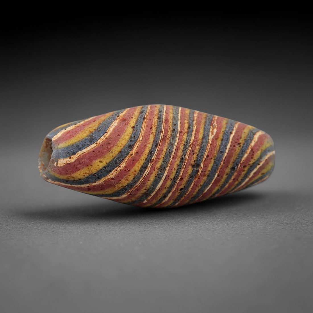 Ancient Islamic Glass Bead - Striped Pattern Amulet Bead, 41FW: This elongated ovoid bead is crafted from layered glass, featuring alternating diagonal stripes of red, yellow, blue, and white that spiral along its length, characteristic of medieval Islamic glasswo