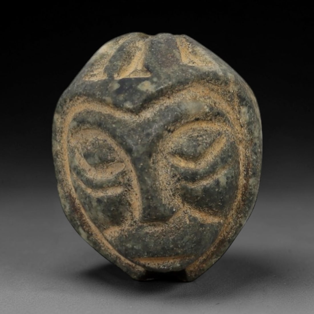 Ancient Stone Face Amulet - Near Eastern Carved Pendant, 65AG: This amulet is carved from a mottled dark green stone with a granular texture and weathered, earthy patina. The form is a stylized human face, with incised linear details defining arched eyebrows, clo