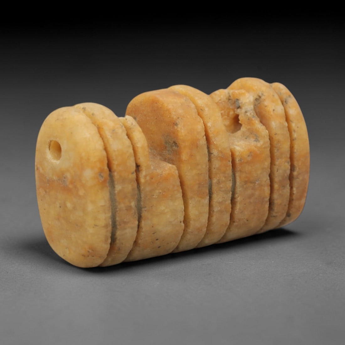 Ancient Stone Ribbed Amulet - Near Eastern Pendant, 75XQ: Carved from a warm ochre stone, this amulet features a segmented, ribbed cylindrical form with a perforated end for suspension, its surface defined by multiple horizontal incised grooves creating stac
